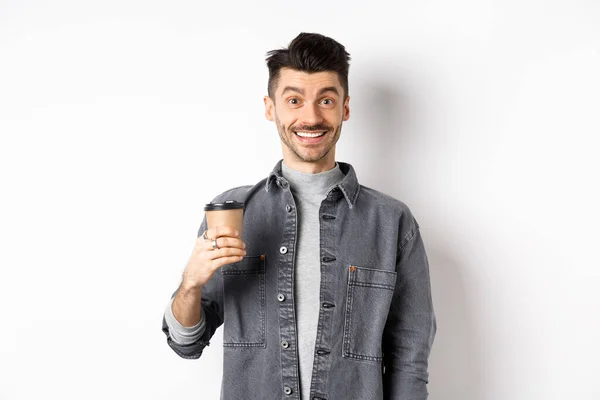 En entusiastisk kjekk mann som holder kaffekopp og smiler, drikker god drink fra kafeteriaen, står opp mot hvit bakgrunn – stockfoto