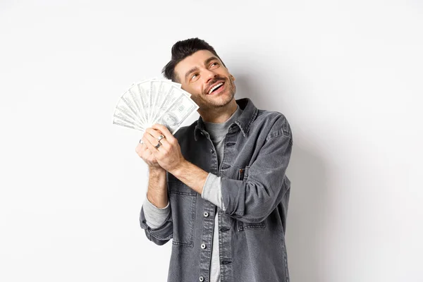 Feliz tipo rico abrazando billetes de dólar y soñando despierto, pensando en ir de compras con dinero, de pie sobre fondo blanco — Foto de Stock