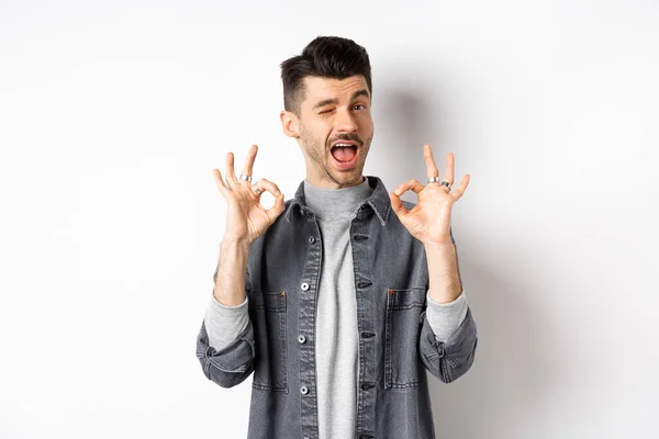 Todo bien. Hombre caucásico guapo mostrando signos OK y sonriendo, guiñando el ojo para alabar lo bueno, recomendando una excelente oferta, de pie sobre fondo blanco —  Fotos de Stock