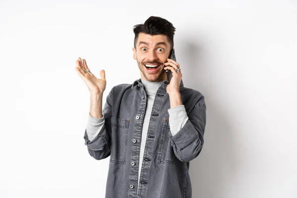 Homem animado falando no celular e ouvir boas notícias, alegrando-se e levantando a mão com sorriso feliz, de pé contra fundo branco — Fotografia de Stock