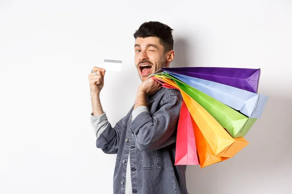 Comprador feliz sosteniendo bolsas de compras sobre el hombro y mostrando tarjeta de crédito de plástico, pagando sin contacto, de pie sobre fondo blanco —  Fotos de Stock