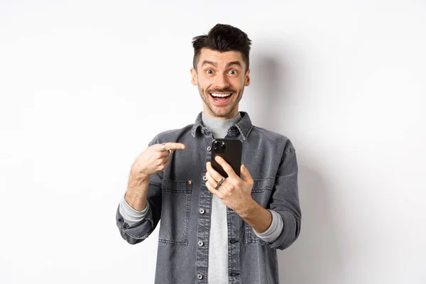 Spännande ung man får goda nyheter på telefon, pekar på smartphone och ler glad, står på vit bakgrund — Stockfoto