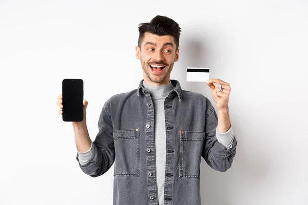 Vzrušený pohledný muž ukazující plastové kreditní karty a prázdné obrazovky smartphone, reklamní aplikace, stojící na bílém pozadí — Stock fotografie