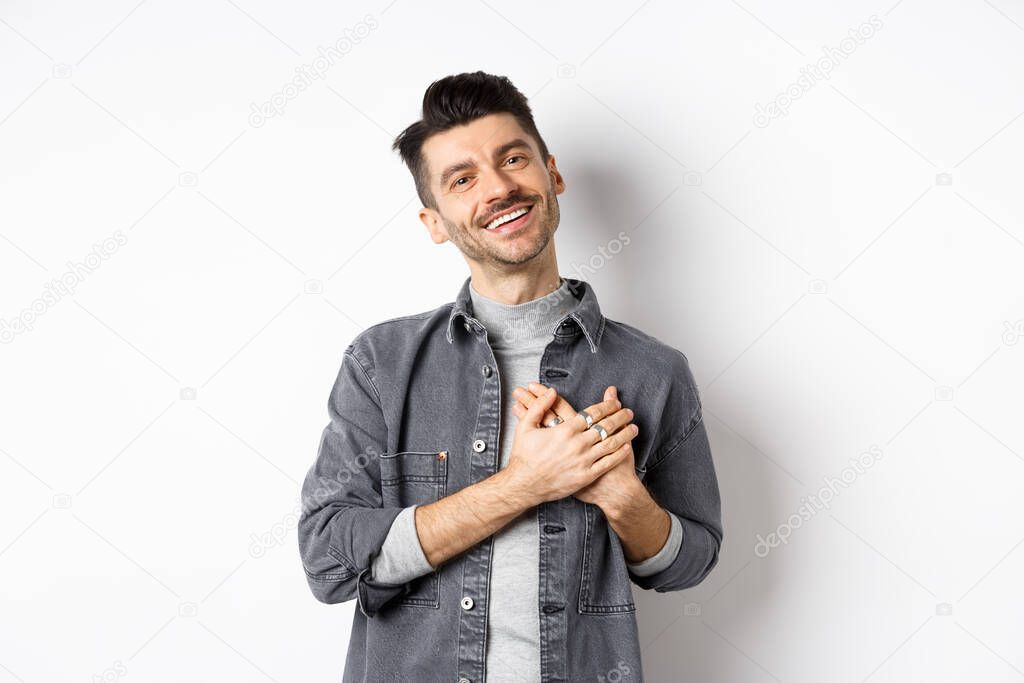 Romantic guy hold hands on heart and smiling, thanking you, looking pleased and grateful, standing on white background
