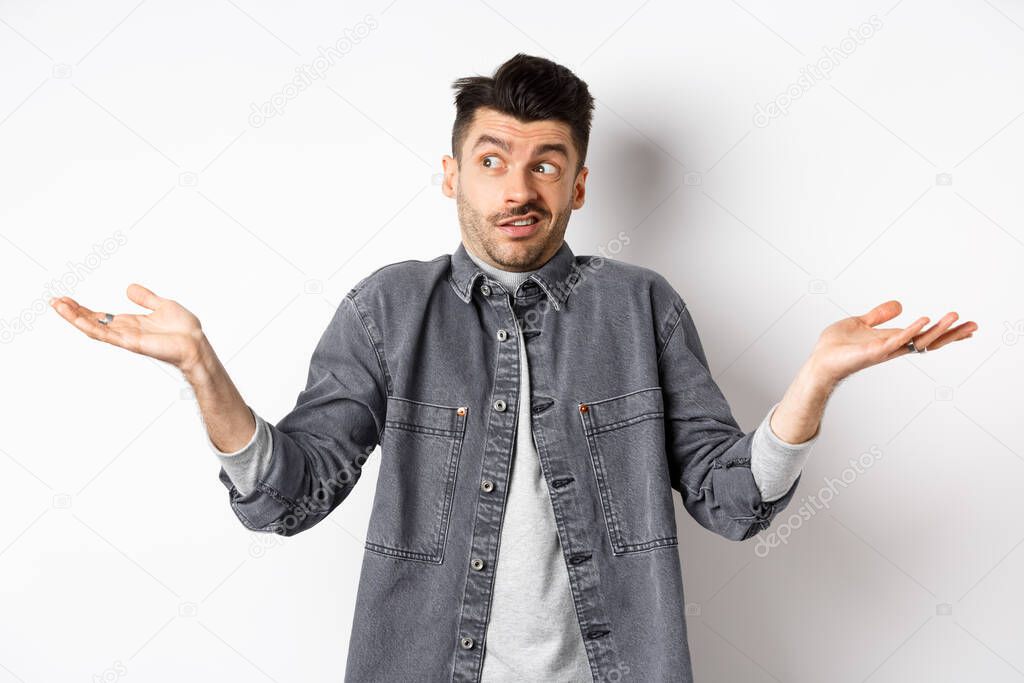 Indecisive young man looking aside and shrugging, know nothing, have no idea, standing clueless and confused against white background