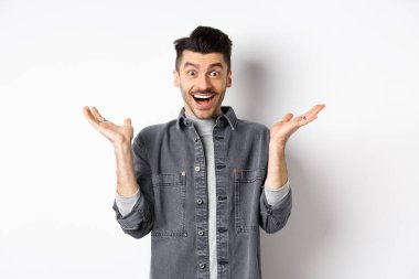 Heyecanlı genç adam sevinçten havalara uçuyor, ellerini yana açıyor ve mutlu görünüyor, şaşkın şaşkın gülüyor, beyaz arka planda duruyor.