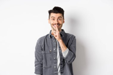Mutlu adam kameraya şşşş yapıyor, parmakları dudaklarında gülümsüyor, sessiz kalmak istiyor, büyük bir sır veriyor ya da sürpriz yapıyor, beyaz arka planda duruyor.