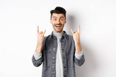 Mutlu ve heyecanlı adam jestle rock yapıyor ve eğleniyor, eğleniyor ve gülümsüyor, beyaz arka planda duruyor.
