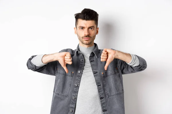 Muy mal. Hombre serio mostrando los pulgares hacia abajo y mirar a la cámara con cara de póker, diciendo que no, desaprueban y no les gusta el producto, de pie insatisfecho sobre el fondo blanco —  Fotos de Stock