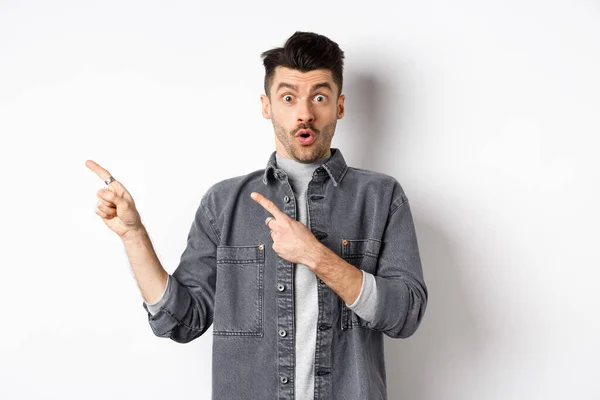 Emocionado hombre apuntando a la izquierda en el logotipo y mirar con incredulidad, diciendo wow a la cámara, echando un vistazo fresco descuento promocional, de pie sobre fondo blanco — Foto de Stock