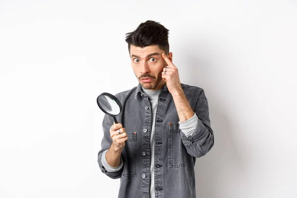 Confused guy found something down with magnifying glass, look puzzled at camera, standing against white background — Stock Photo, Image