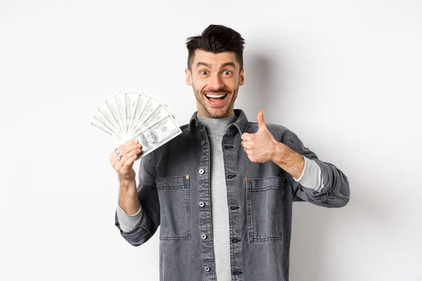 Obraz podekscytowanego człowieka trzymającego dolary i pokazującego kciuki w górę szczęśliwą twarzą, zarabiającego pieniądze, stojącego na białym tle — Zdjęcie stockowe