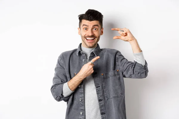 Opgewonden man met klein formaat, wijzende vinger naar klein ding en glimlachend, staande tegen een witte achtergrond — Stockfoto