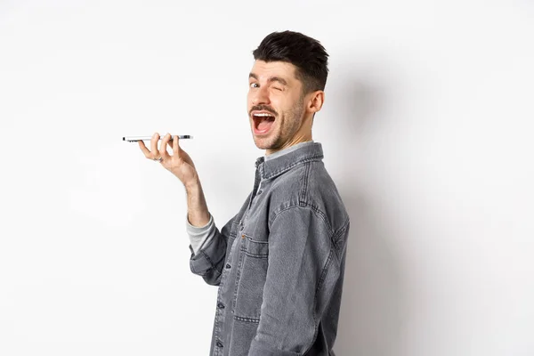 Happy guy knipperen op het kijken naar de camera tijdens het gebruik van voice translator app of praten op de luidspreker, houden telefoon dicht bij de mond, staan op een witte achtergrond — Stockfoto