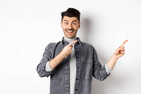 Kijk hier. Gelukkige stijlvolle man glimlachend, wijzende vingers recht naar lege ruimte, tonen coole promo aanbod, staande in denim jas op witte achtergrond — Stockfoto