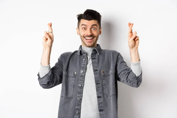 Jovem otimista do sexo masculino esperando o sonho se tornar realidade, fazendo desejo com os dedos cruzados, sorrindo e esperando boas notícias, de pé sobre fundo branco — Fotografia de Stock