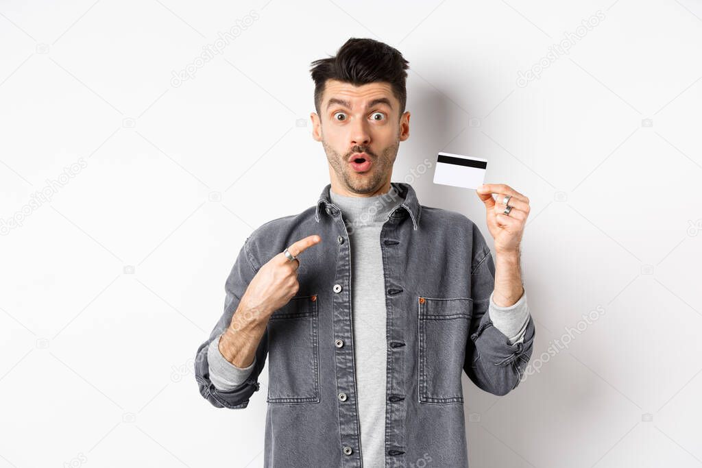 Wow look here. Excited guy pointing at plastic credit card and look amazed with awesome deal, standing against white background