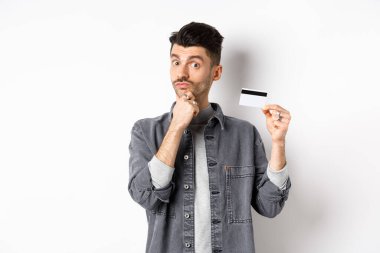 Düşünceli bir adam plastik kredi kartı tutuyor ve düşünüyor, kameraya bakıyor, seçim yaparken çenesine dokunuyor, beyaz arka planda duruyor.