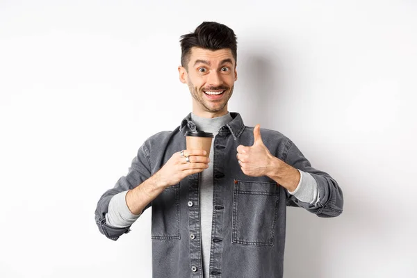 Muy buena bebida. Sonriendo chico elegante beber café y mostrando los pulgares hacia arriba, tienda de café de alabanza, de pie satisfecho en el fondo blanco —  Fotos de Stock