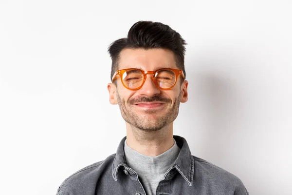 Close-up de alegre sorridente homem fechar os olhos, sentindo felicidade e alegria, satisfeito com algo bom, vestindo óculos da moda, fundo branco — Fotografia de Stock