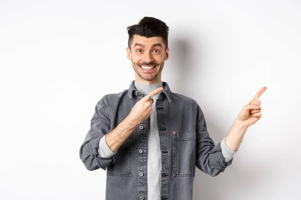 Vrolijke stijlvolle man met snor en witte glimlach, wijzende vingers naar rechts, uitnodigend om hier te kijken, staande op witte achtergrond — Stockfoto