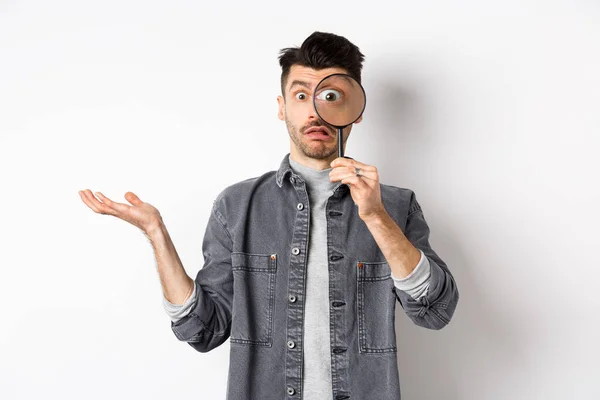 Confused man look through magnifying glass and shrugging clueless, standing on white background — Stock Photo, Image