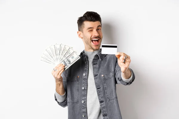 Imagen de tipo guapo sosteniendo billetes de dólar pero sugiriendo utilizar tarjeta de crédito de plástico, sonriendo amigable a la cámara, de pie sobre fondo blanco —  Fotos de Stock