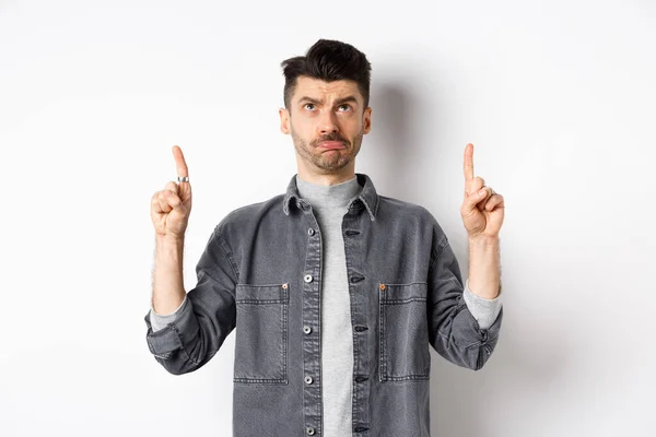 Sad frowning guy pointing and looking up, standing hesitant on white background — Stock Photo, Image