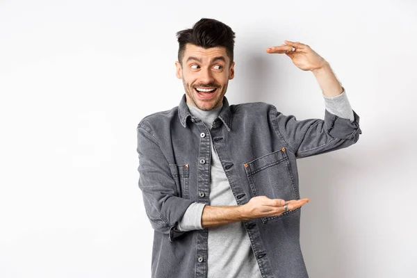 Opgewonden man kijken naar logo en tonen grote maat ding, vormgeven van grote object en glimlachend verbaasd, staande in denim jas op witte achtergrond — Stockfoto