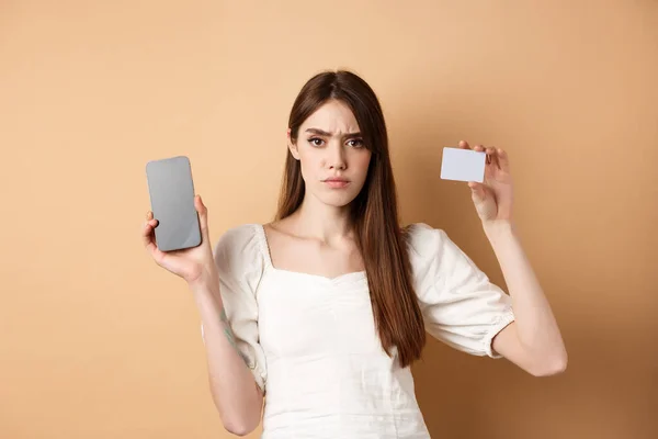 Menina triste e decepcionada mostrando tela de smartphone vazio e cartão de crédito de plástico, reclamando e franzindo a testa, de pé sobre fundo bege — Fotografia de Stock