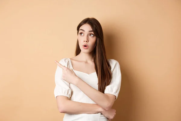 Young woman in dress looking and pointing at upper left corner wondered, saying wow, checking out advertisement with intrigued face, beige background — Stock Photo, Image