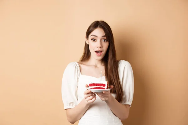 Garota de aniversário animado olhando espantado, segurando bolo com vela, fazendo b-dia desejo, de pé sobre fundo bege — Fotografia de Stock