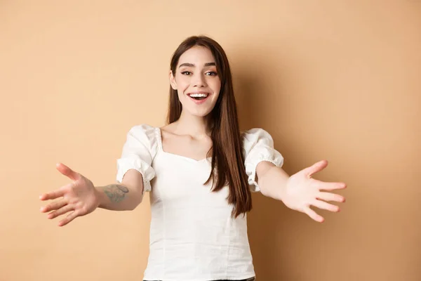 Retrato de menina franca amigável chegar para abraço, bem-vindo amigo e sorrindo, estender as mãos lateralmente em gesto de saudação, recebendo algo, fundo bege — Fotografia de Stock