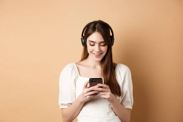 Mujer sonriente mensaje de texto en el teléfono y escuchar música en los auriculares inalámbricos, viendo vídeo en el teléfono inteligente, fondo beige —  Fotos de Stock