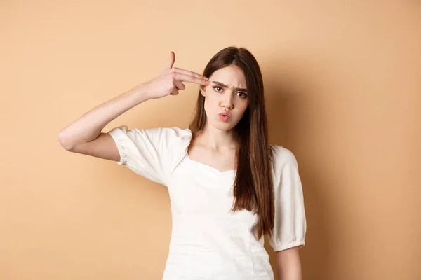 Just kill me. Annoyed and distressed young woman making finger gun sign on ver head and frowning bothered, standing pissed-off on beige background — Foto Stock