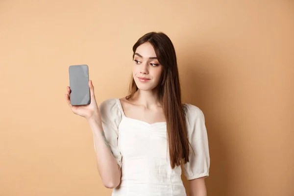 Atraktivní mladá žena ukazuje prázdný displej smartphonu, usmívá se a dívá se stranou, stojí na béžovém pozadí — Stock fotografie