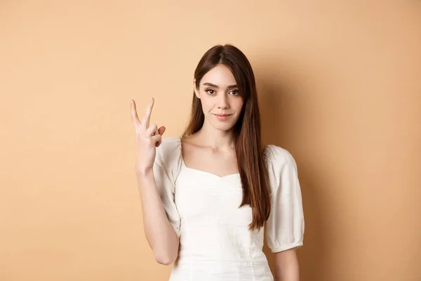 Ελκυστική νεαρή γυναίκα δείχνουν τα δάχτυλα νούμερο δύο, χαμογελώντας και αναζητούν αυτοπεποίθηση, στέκεται σε μπεζ φόντο — Φωτογραφία Αρχείου