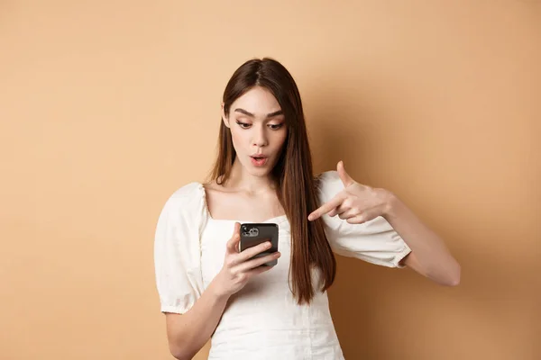 Vzrušující zprávy po telefonu. Překvapená roztomilá žena čtení online promo na obrazovce, ukazuje na smartphone a říct wow ohromen, stojící na béžové pozadí — Stock fotografie
