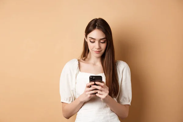 Hezká žena smskování na smartphone mobil, čtení obrazovky a usmívání, stojí na béžové pozadí — Stock fotografie