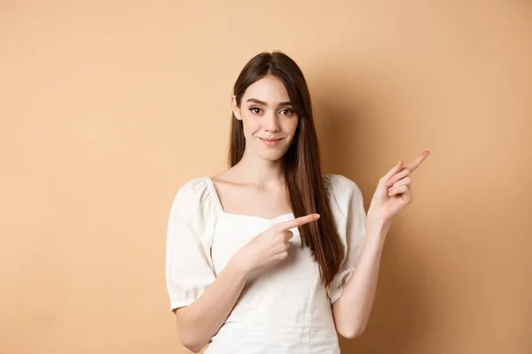 Mulher bonita no vestido apontando os dedos direito, mostrando logotipo e olhando concurso para a câmera, fundo bege — Fotografia de Stock