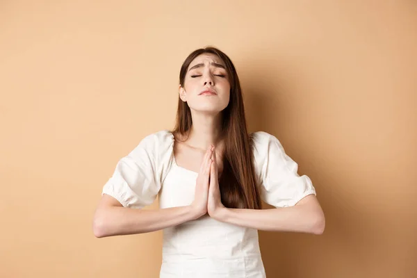 Femme pleine d'espoir fermer les yeux et prier Dieu, tenant la main dans le geste de mendicité et faire le vœu, dire s'il vous plaît, debout sur fond beige — Photo