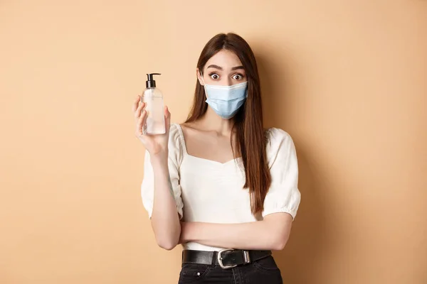 Salgın ve sağlık sigortası konsepti. Yüz maskesi takmış, el dezenfektanı şişesini gösteren heyecanlı bir kız, kameraya bakın, bej arka planda duruyor. — Stok fotoğraf