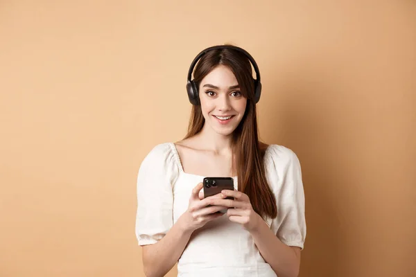 Chica bonita en auriculares sonriendo a la cámara, escuchando música y utilizando la aplicación del teléfono móvil, fondo beige — Foto de Stock