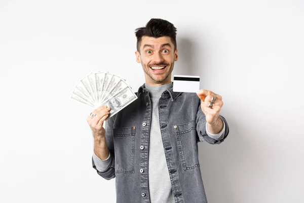 Imagen de tipo guapo sosteniendo billetes de dólar pero sugiriendo utilizar tarjeta de crédito de plástico, sonriendo amigable a la cámara, de pie sobre fondo blanco — Foto de Stock
