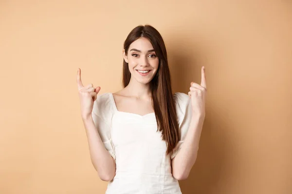 Atractiva mujer caucásica en vestido señalando los dedos hacia arriba, sonriendo y mostrando pancarta promocional, de pie sobre fondo beige — Foto de Stock