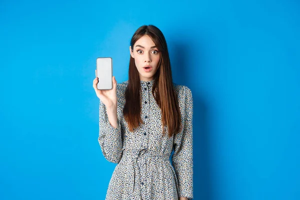 Возбужденная молодая женщина в платье, задыхаясь удивлен, показывая пустой экран смартфона и смотреть впечатлен камерой, синий фон — стоковое фото