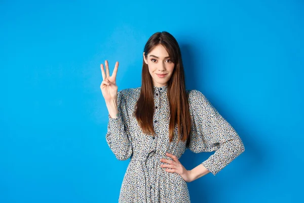 Menina bonita nova no vestido com cabelo longo natural, mostrando o número três com os dedos e sorrindo, de pé contra o fundo azul — Fotografia de Stock