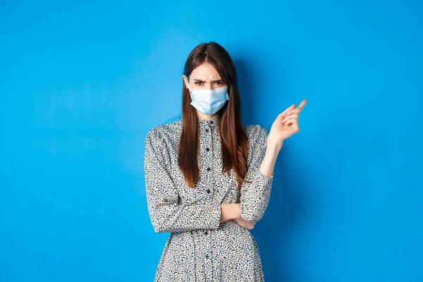 Covid-19, concept de distance sociale et de soins de santé. Jeune femme en colère dans le masque médical fronçant les sourcils, pointant droit sur la mauvaise chose, condamner les non-masqueurs, fond bleu — Photo