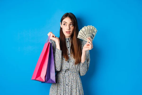 Shopping. Pretty woman thinking what to buy, looking left at logo, holding paper shop bags and money, standing on blue background — Stock Photo, Image