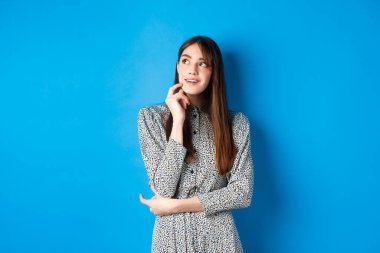 Yaratıcı genç bir kadın hayal ediyor ve gülümsüyor, sol üst logoya bakıyor düşünceli, bir fikri var, mavi arka planda duruyor.